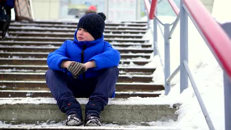 Junge-Teenager-in-blau-Daunenjacke-verloren-in-der-Stadt.-Er-sitzt-auf-einer-kalten-Treppe,-schaut-sich-um.