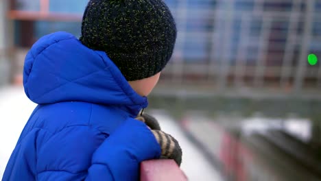 Boy-teenager-in-blue-down-jacket-lost-in-the-city.-He-is-at-the-train-station,-looking-down.