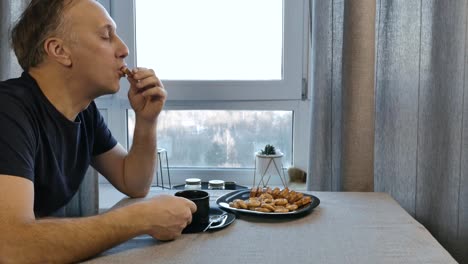 The-man-has-breakfast-in-the-morning-at-home-in-the-kitchen,-and-he-is-in-a-good-mood.-He-drinks-coffee-and-eating-homemade-cookies.