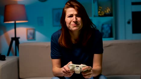 La-chica-triste-pierde-el-videojuego-en-casa-por-la-noche.-Mujer-jugadora-excitada-sentada-en-un-sofá,-jugando-y-perdiendo-en-videojuegos-en-una-consola,-usando-un-mando-inalámbrico.-Habitación-acogedora,-ambiente-casero-encantador