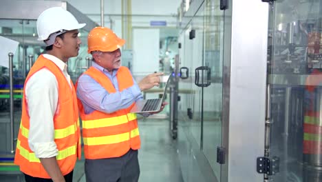 Senior-quality-inspector-teaching-junior-team-member-in-factory.-Chinese-old-male-with-his-young-team-discussing-quality-issue-with-bottle-production-line-in-background.