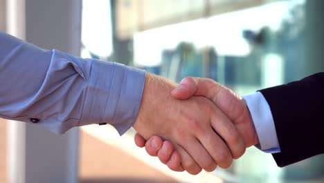 Close-up-of-two-successful-businessmen-greeting-each-other-in-urban-environment.-Young-colleagues-meeting-and-shaking-hands-near-office-building.-Handshake-of-business-partners-outdoor.-Side-view