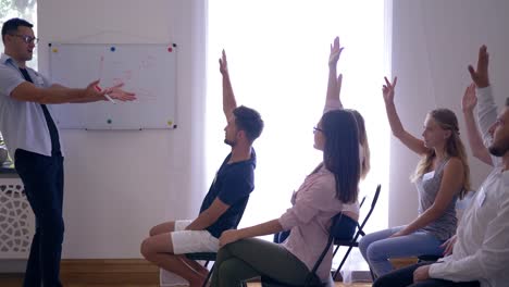 interactive-training-on-seminar,-coach-gives-presentation-of-new-project-and-listens-to-colleagues-raising-hands-up-in-room