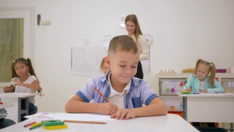 Kinder-im-Grundschulalter-zeichnen-auf-Papier-mit-Farbstiften-an-einem-Schreibtisch-mit-junger-Lehrer-in-helle-Klassenzimmer