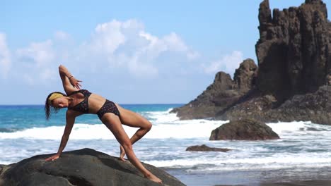 Mujer-haciendo-fitness-en-la-playa-con-arena-negra-volcánica-en-un-traje-de-baño-sentado-en-una-roca-en-las-montañas-de-lava.-El-concepto-de-estilo-de-vida-saludable-y-la-recreación-en-las-islas