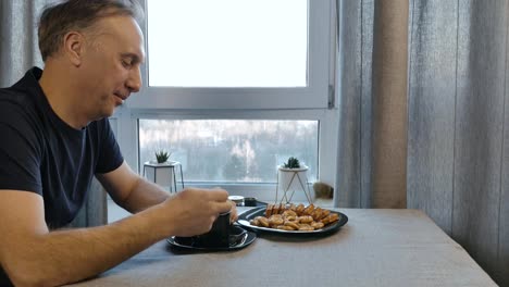 Un-hombre-de-mediana-edad-en-la-mañana-tomando-café-y-comiendo-galletas-hechas-en-casa.-Él-se-sienta-en-casa-en-la-cocina-y-él-está-de-buen-humor.