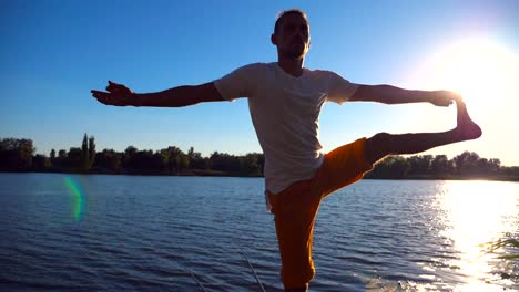 Young-man-standing-at-yoga-pose-on-wooden-jetty-at-lake.-Athlete-balancing-on-one-leg-at-nature.-Sporty-guy-doing-stretch-exercise-outdoor.-Concept-of-healthy-active-lifestyle.-Slow-motion-Close-up