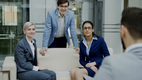 Team-of-cheerful-business-colleagues-talking-and-discussing-each-other-while-having-break-sitting-on-sofa-in-modern-office-hall