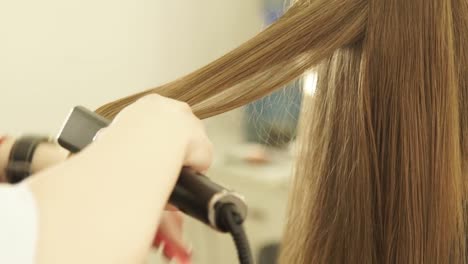 Hairstylist-combing-long-hair-and-straightening-with-hair-tongs-in-hairdressing-salon.-Close-up-female-hairdresser-straightening-hair-during-hairstyling-in-beauty-salon