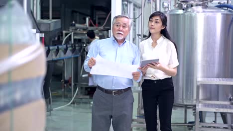 Factory-owner-inspecting-his-production-line.-Asian-senior-man-with-beautiful-asian-female-personal-assistant-talking-inside-factory.