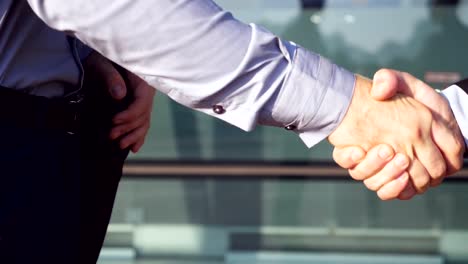 Two-confident-businessmen-shaking-hands-after-successful-deal-near-office-building.-Young-colleagues-congratulating-each-other-in-urban-environment.-Handshake-of-business-partners.-Slow-motion