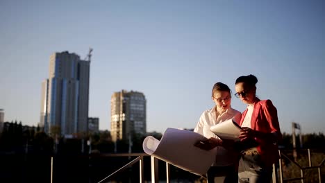 Dos-mujeres-con-los-planos-y-documentación-técnica-en-el-paseo-marítimo-en-el-fondo-de-los-rascacielos-en-construcción-para-discutir-los-riesgos-y-la-rentabilidad-de-la-construcción-de-un-nuevo-centro-de-negocios.