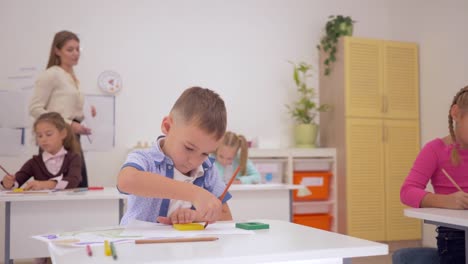 Grundschule,-zeichnen-Schüler-auf-Papier-mit-Farbstiften-sitzen-an-einem-Schreibtisch-in-helle-Klassenzimmer-mit-jungen-Lehrer