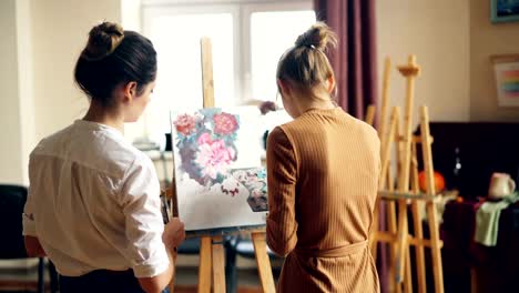 Back-view-of-art-student-painting-flowers-on-canvas-and-her-helpful-teacher-standing-nearby-and-checking-her-work.-Visual-arts-and-education-concept.