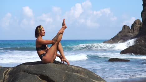 A-young-girl-in-a-bikini-balancing-sitting-on-a-stone-and-doing-exercises-for-the-abdominal-muscles-alternately-lifting-her-legs.-Training-on-the-background-of-the-ocean-on-the-black-sand-beach