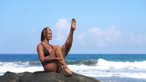 Woman-doing-fitness-on-the-beach-with-volcanic-black-sand-in-a-swimsuit-sitting-on-a-rock-around-the-mountains-of-lava.-The-concept-of-healthy-lifestyle-and-recreation-on-the-Islands