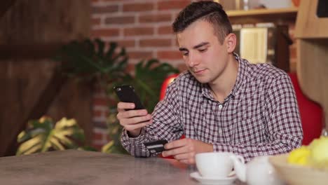 Handsome-man-making-purchase-online,-typing-credit-card-details-on-black-smartphone-in-the-modern-kitchen