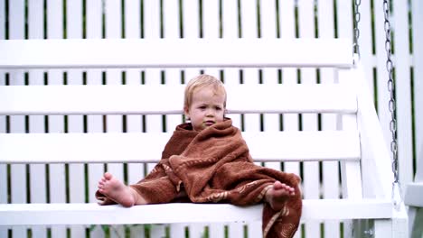 a-little-one-year-old-girl,-wrapped-in-a-towel,-with-a-dirty-face,-lonely-sitting-on-a-swing-in-the-garden,-in-the-summer.-She-has-a-sad-look.-She-wants-to-sleep