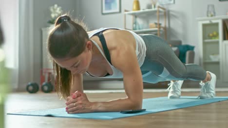 Fuerte-hermosa-chica-Fitness-en-ropa-de-entrenamiento-atlético-es-haciendo-una-tabla-ejercicio-mientras-utiliza-un-cronómetro-en-su-teléfono.-Ella-está-entrenando-en-su-casa-en-su-sala-de-estar-con-Interior-acogedor.