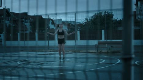 Hermosa-chica-de-aptitud-energética-Top-deportivo-negro-y-pantalones-cortos-es-saltan/salto-de-cuerda.-Ella-está-haciendo-un-entrenamiento-en-una-cancha-de-baloncesto-al-aire-libre-cercado.-Vista-desde-detrás-de-la-valla.-Noche-después-de-la-lluvia.