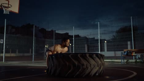 Fuerte-ajuste-joven-sin-camisa-hombre-musculoso-es-haciendo-ejercicios-en-una-cancha-de-baloncesto-al-aire-libre-cercado.-Él-está-cambiando-un-neumático-pesado-grande-en-una-tarde-después-de-la-lluvia-en-una-zona-de-barrio-residencial.