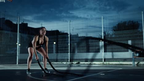 Adecuación-energética-hermosa-chica-haciendo-ejercicios-con-cuerdas-de-batalla.-Ella-está-haciendo-un-entrenamiento-en-una-cancha-de-baloncesto-al-aire-libre-cercado.-Imágenes-de-noche-después-de-la-lluvia-en-una-zona-de-barrio-residencial.