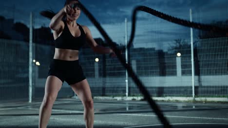 Adecuación-energética-hermosa-chica-haciendo-ejercicios-con-cuerdas-de-batalla.-Ella-está-haciendo-un-entrenamiento-en-una-cancha-de-baloncesto-al-aire-libre-cercado.-Imágenes-de-noche-después-de-la-lluvia-en-una-zona-de-barrio-residencial.