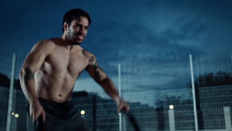 Strong-Muscular-Fit-Shirtless-Young-Man-is-Doing-Exercises-with-Battle-Ropes.-He-is-Doing-a-Workout-in-a-Fenced-Outdoor-Basketball-Court.-Evening-Footage-After-Rain-in-a-Residential-Neighborhood-Area.