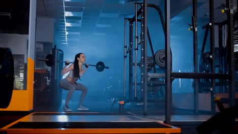 Girl-crouches-with-a-barbell-behind-her-back-in-the-gym.-Smoke,-darkness