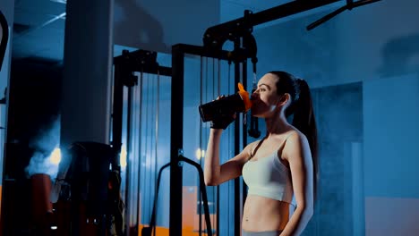 Mädchen-steht-am-Simulator,-müde-nach-dem-Training.-Trinken-Sie-aus-einem-Boston-Shaker.-Rauch,-Trübsinn,-Fitness-Studio