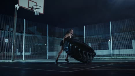 Schöne-energetische-Fitness-Mädchen-ist-dabei-Übungen-in-einem-eingezäunten-Basketballfeld-im-Freien.-Sie-ist-einen-große-schwere-Reifen-in-einer-nebligen-Nacht-nach-Regen-in-einer-Wohngegend-Gegend-spiegeln.