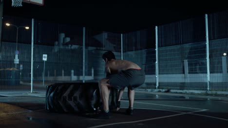 Backshot-de-un-fuerte-ajuste-joven-sin-camisa-hombre-haciendo-ejercicios-en-una-cancha-de-baloncesto-al-aire-libre-cercado.-Él-está-cambiando-un-neumático-pesado-grande-en-una-noche-después-de-la-lluvia-en-una-zona-de-barrio-residencial.