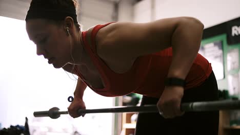 Un-ajuste-mujer-joven-levantar-la-barra-para-pesas-en-un-pequeño-gimnasio-mientras-escuchando-música