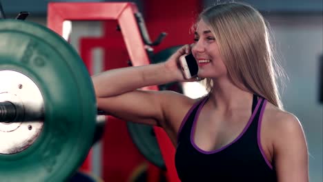 Lächelnde-Pass-Frau-im-Gespräch-am-Telefon-im-Fitnessstudio-Gewichtsraum