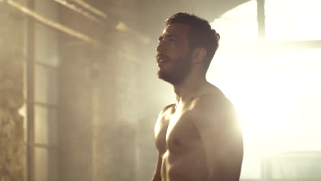 Handsome-Shirtless-Man-with-Naked-Muscular-Torso-with-Visible-Six-Pack-Stands-Resting-after-Bodybuilding-Exercise,-He-Wipes-Sweat-from-His-Forehead.-He's-in-the-Middle-of-Abandoned-Factory-Building.