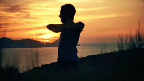 Silhouette-of-a-man-stretching-on-top-of-a-mountain-on-an-island-at-sunset-with-the-sea-on-the-background