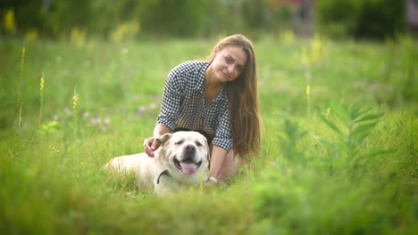 mujer-bonita-es-acariciando-su-labrador-blanco-grande-y-mirando-en-la-cámara-en-medio-de-un-campo-verde,-verde-hierba