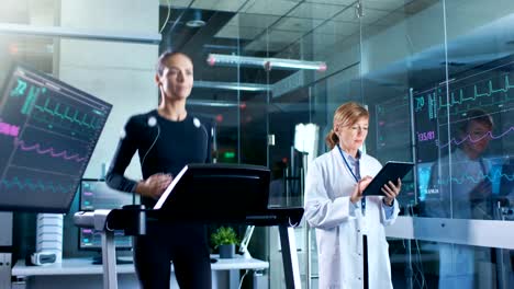 Woman-Athlete-Runs-on-a-Treadmill--with-Electrodes-Attached-to-Her-Body-while-Scientist-Holding-Tablet-Computer-Supervises-whole-Process.-In-the-Background-Laboratory-with-Monitors-Showing-EKG-Readings.