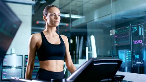 Hermosa-mujer-atleta-llevar-sujetador-de-deporte,-camina-en-una-cinta-de-correr-en-un-laboratorio-de-ciencia-de-deportes.-En-el-fondo-monitores-ver-electrocardiograma-datos-y-datos-de-salud.