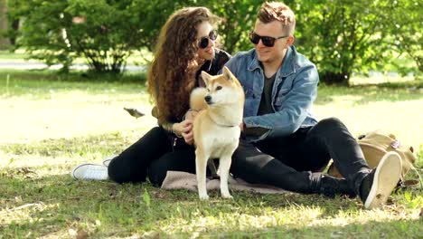 Husband-and-wife-happy-couple-are-fussing-their-pet-shiba-inu-puppy-sitting-on-grass-in-park-and-talking.-Conversation,-animals-and-modern-lifestyle-concept.