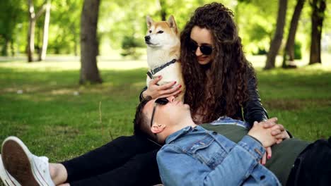 Cheerful-young-man-is-lying-on-the-grass-in-the-park-with-his-head-on-his-wife's-legs-while-attractive-smiling-woman-is-talking-to-him-and-caressing-well-bred-dog.