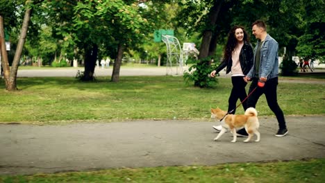 Cheerful-young-couple-man-and-woman-are-walking-the-dog-in-the-park-near-sports-ground,-people-are-holding-hands,-guy-is-leading-the-animal.-Urban-life-and-pets-concept.