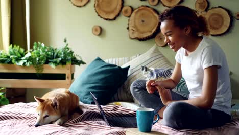 Happy-girl-is-skyping-talking-online-with-friends-gesturing-and-looking-at-laptop-screen-sitting-on-bed-at-home-with-puppy-lying-beside-her.-Communication-and-technology-concept.