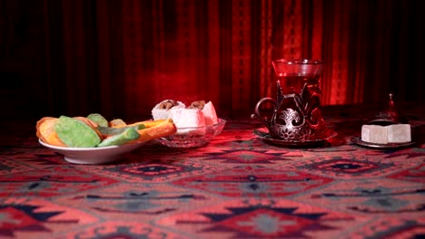 Arabian-tea-in-vintage-glass-with-eastern-snacks-on-a-carpet.-Eastern-tea-ceremony-on-dark-background-with-lights-and-smoke.-Empty-space.-Selective-focus.-Slider-shot