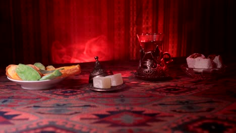 Arabian-tea-in-vintage-glass-with-eastern-snacks-on-a-carpet.-Eastern-tea-ceremony-on-dark-background-with-lights-and-smoke.-Empty-space.-Selective-focus.-Slider-shot