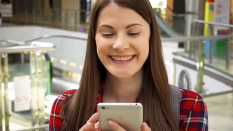 Young-beautiful-woman-standing-in-shopping-mall-smiling.-Using-her-smartphone,-talking-with-friends