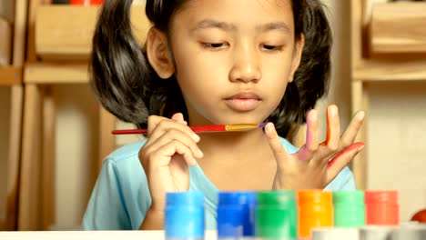 The-little-girl-is-using-a-red-paintbrush-dipped-in-a-colored-bottle-to-paint-on-her-finger.