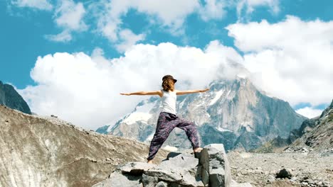 Joven-realiza-una-asana-básica-de-Guerrero-en-el-yoga-contra-el-telón-de-fondo-de-una-montaña-cubierto-de-nieve-en-una-excursión.-Niña-hace-gimnasia-y-gimnasia-en-aire-fresco-en-una-caminata-en-la-naturaleza