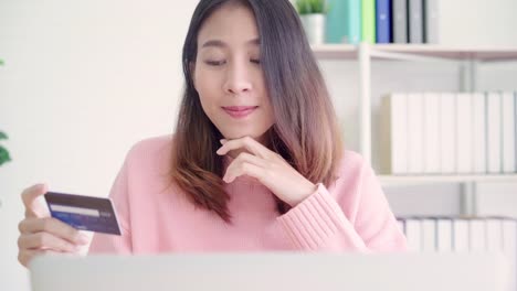 Beautiful-Asian-woman-using-computer-or-laptop-buying-online-shopping-by-credit-card-while-wear-sweater-sitting-on-desk-in-living-room-at-home.-Lifestyle-woman-at-home-concept.