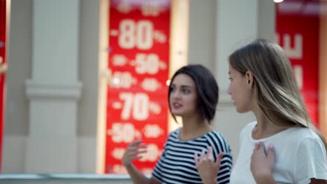 Two-young-women-spending-time-in-shopping-mall-in-sales-period.-Female-friends-walking-and-talking-during-Black-Friday-shopping